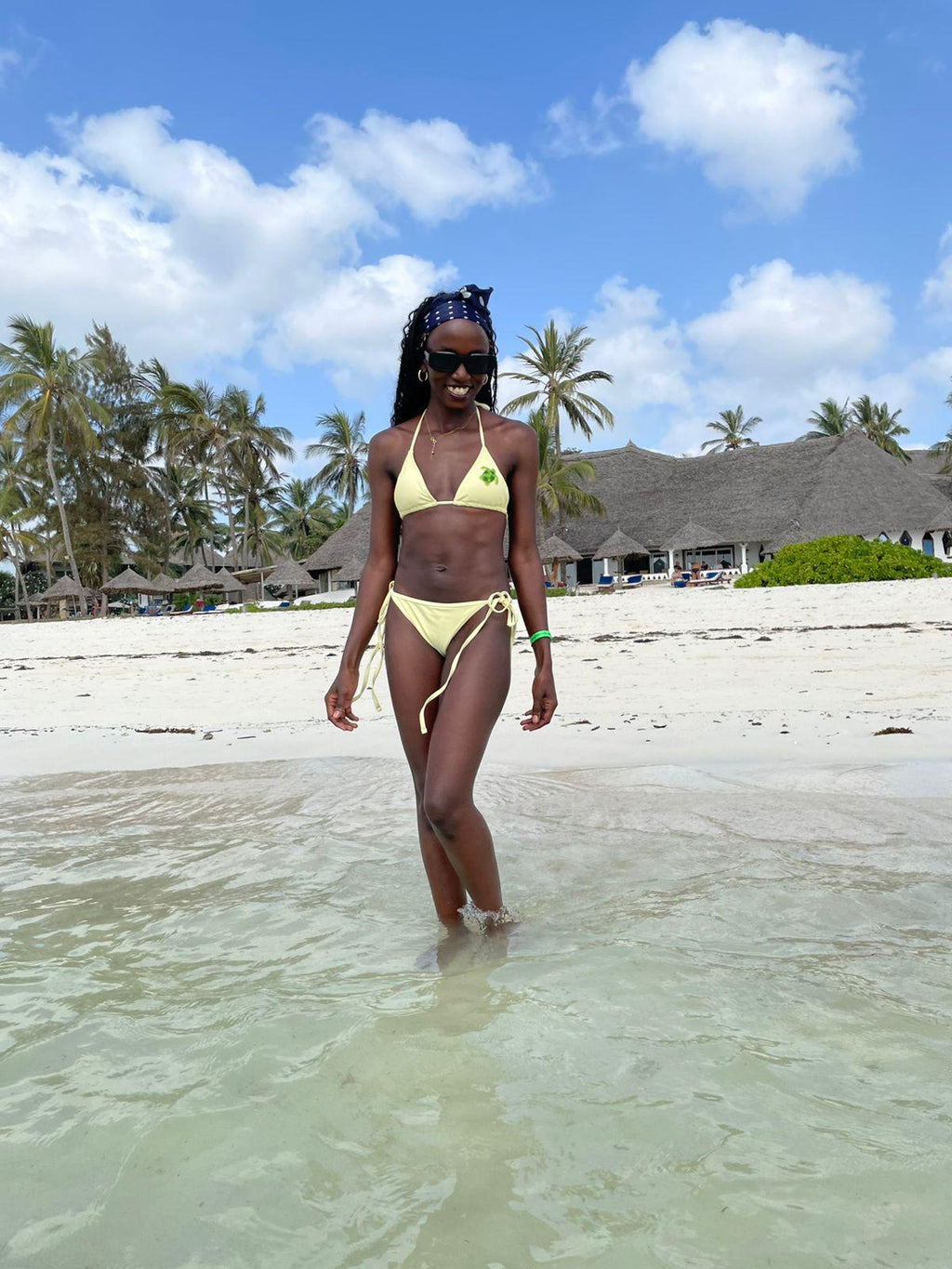 A woman wearing a light yellow bikini walks through shallow ocean water on a sandy beach with palm trees and resort buildings in the background.